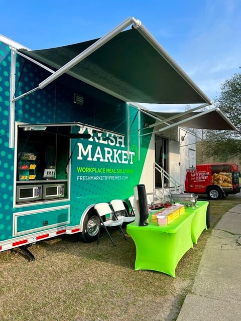 Green and teal food truck with Fresh Market signage and extended awning, parked on a street with bright green table covering